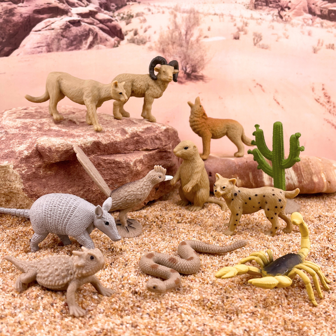 Desert themed animal figurines placed in a DIY desert diorama with sand and rocks and a picture of a desert in the background.