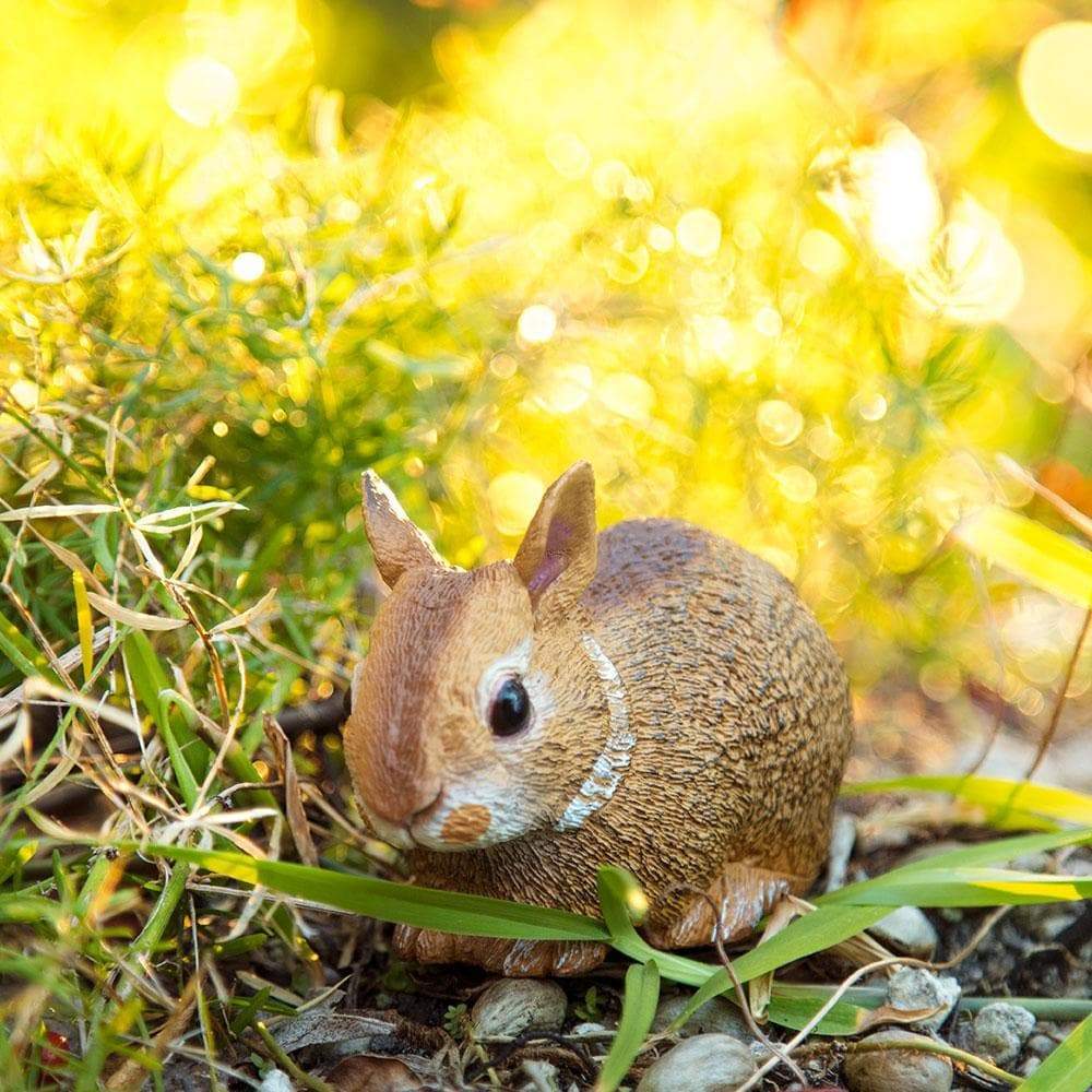 A small, brown rabbit figurine sits on the ground among green grass and plants, with sunlight creating a warm, golden bokeh effect in the background.