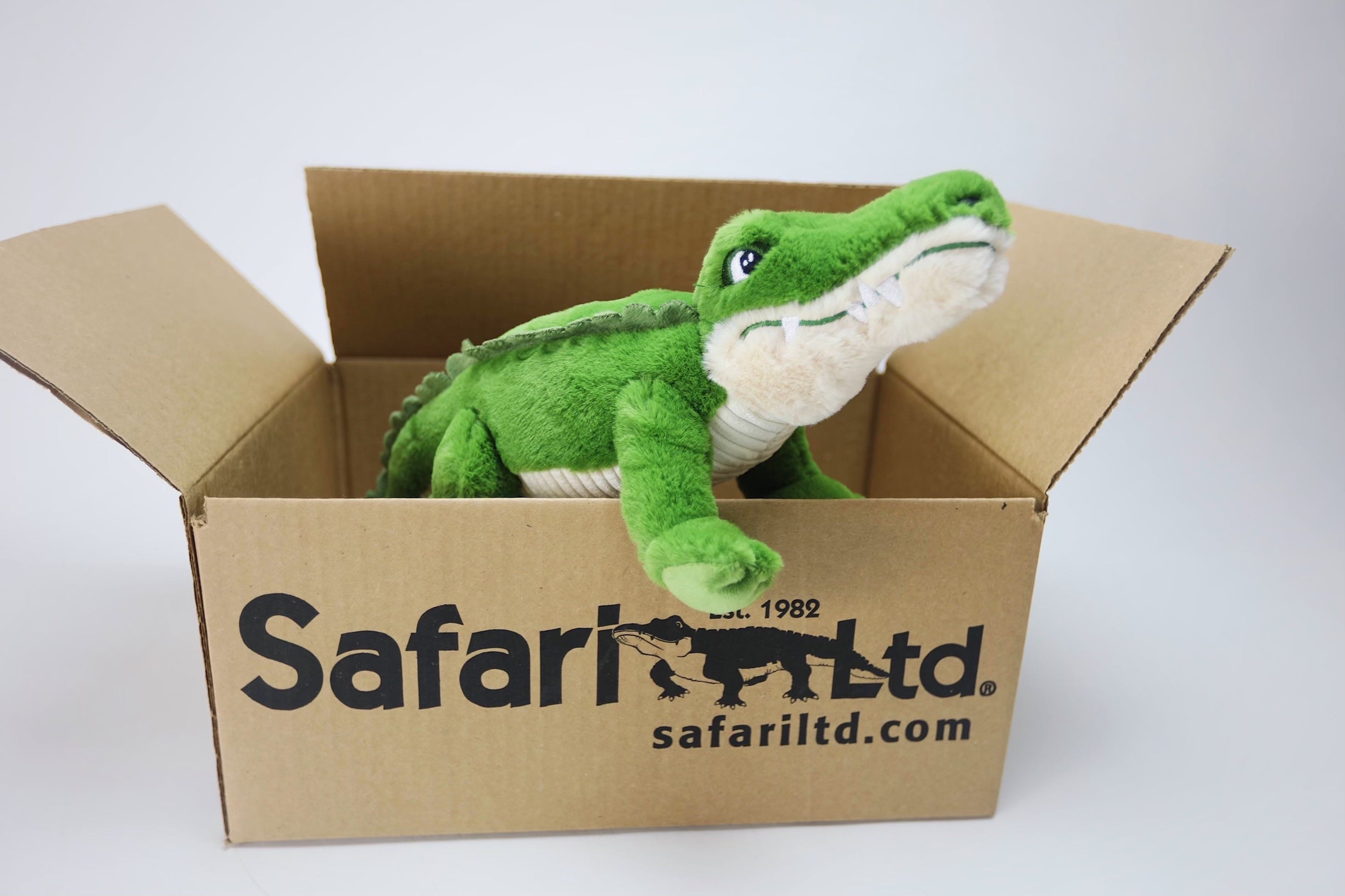 A green plush crocodile toy sits inside a cardboard box with the Safari Ltd. logo, featuring a crocodile graphic and the website safariltd.com, on a plain white background.