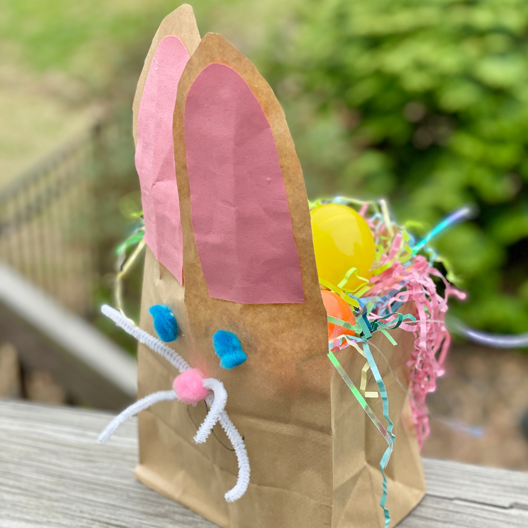 A brown paper bag decorated as a bunny, with pink paper ears, blue pom-pom eyes, a pink pom-pom nose, and white pipe cleaner whiskers, filled with colorful plastic eggs and shredded paper grass.