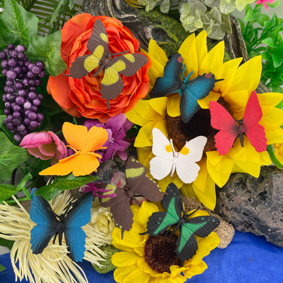 Colorful artificial butterflies rest on bright artificial flowers, including sunflowers, an orange rose, and purple grapes, surrounded by green leaves and plants. The scene is vivid and decorative.