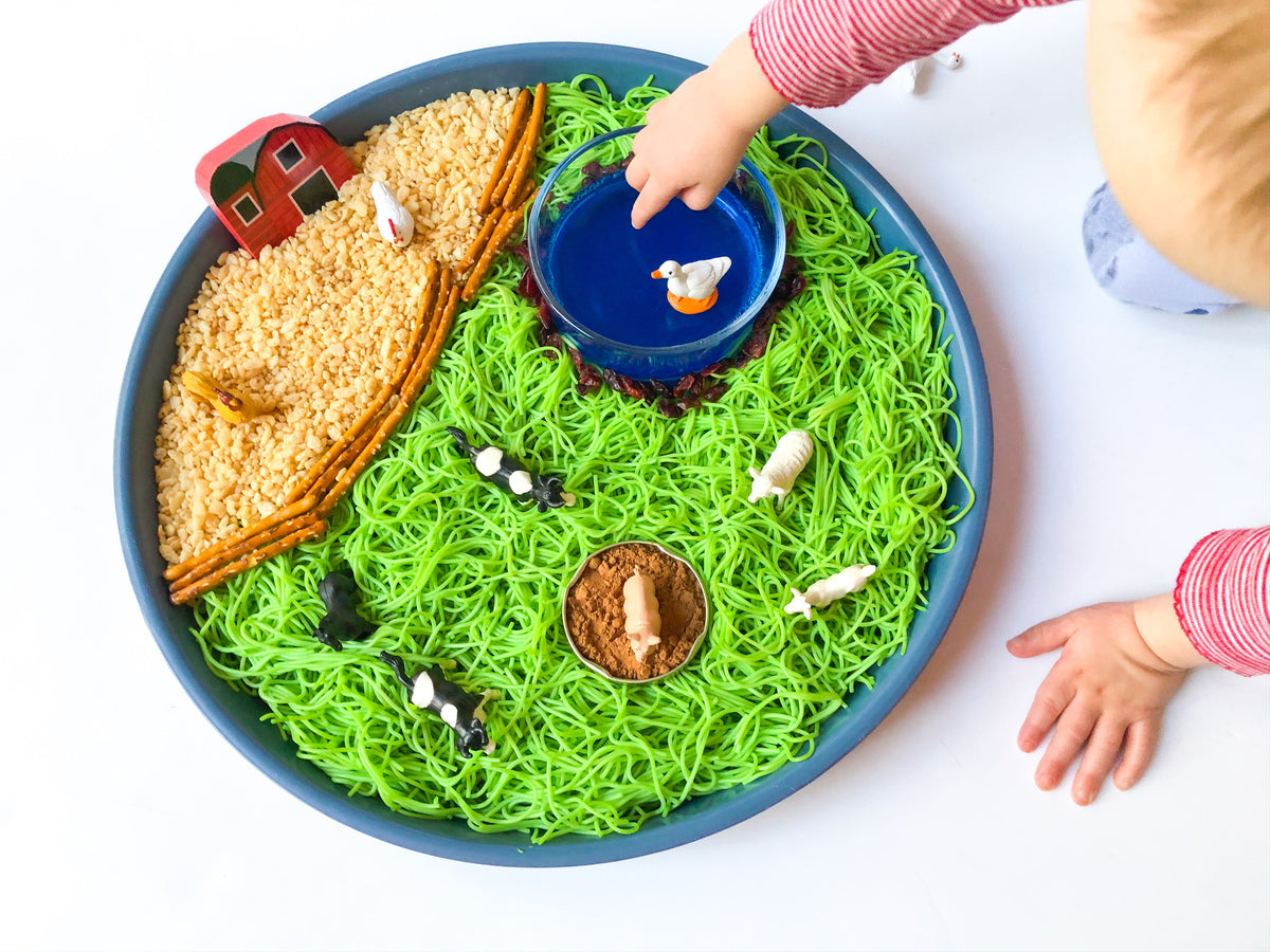 A childs hands reach toward a farm-themed sensory play tray with green noodles as grass, brown rice as a field, animal figurines, a toy barn, and a blue bowl representing a pond.