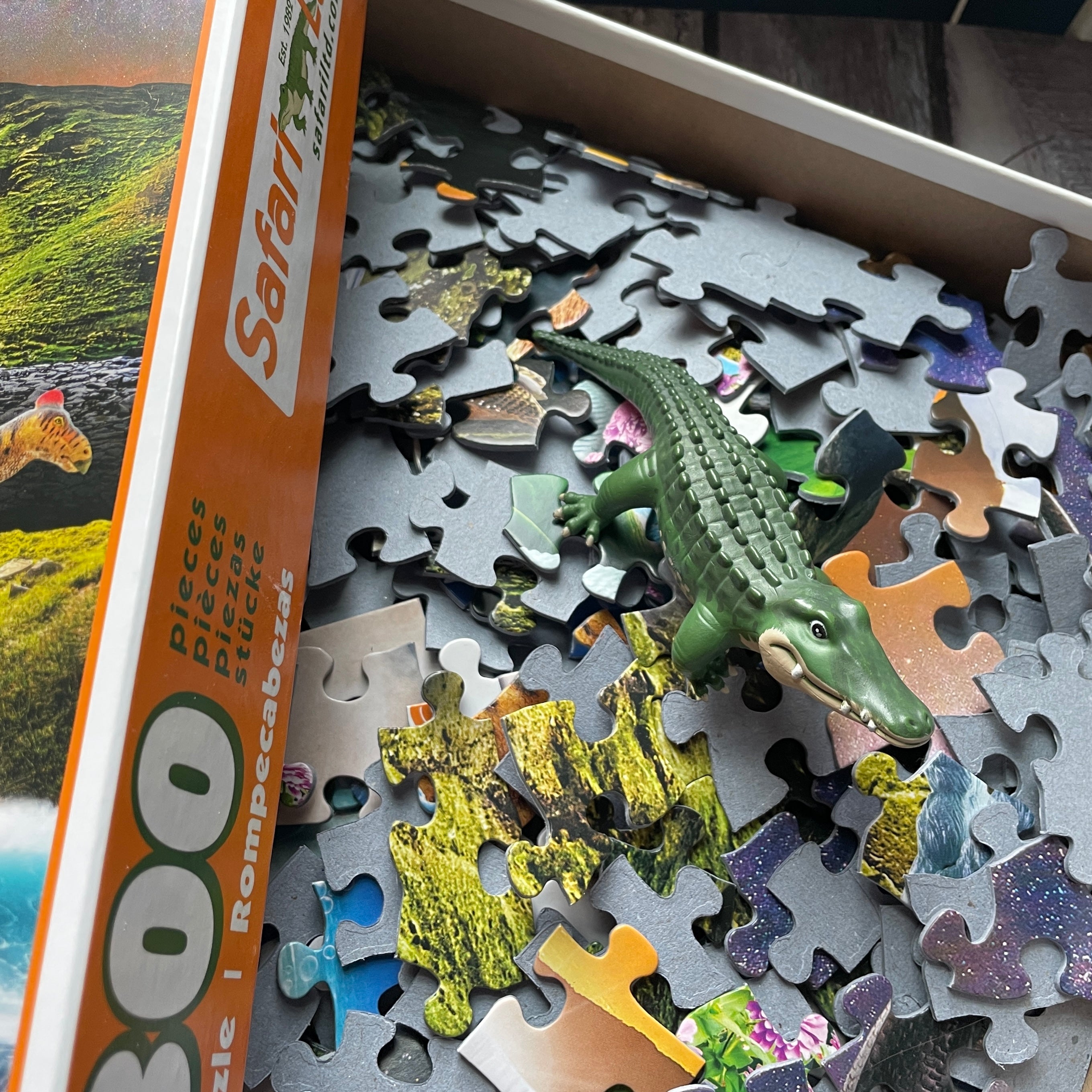 A close-up of an open puzzle box labeled Safari with scattered jigsaw pieces inside and a small green toy alligator resting on top of the pieces.