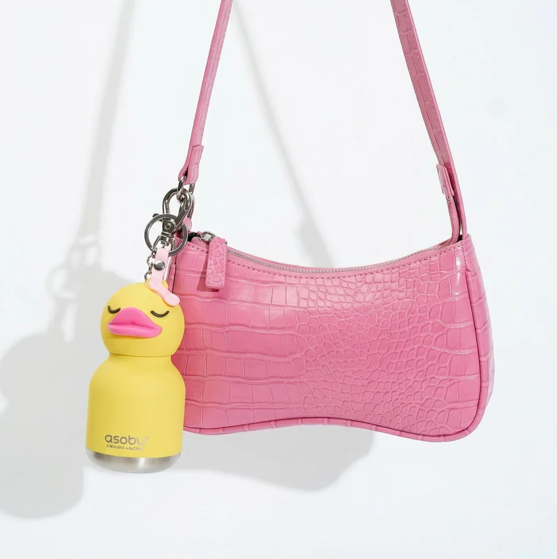 A pink crocodile-patterned handbag is shown with an Asobu Bottle/AdnArt Inc Mini Duck Best Bottle Vacuum Flask, featuring an animal keychain shaped like a yellow duck with closed eyes and a pink bow, photographed against a plain white background.