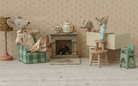 Maileg Twins, Baby Mice In Matchbox by Maileg are featured in a tiny living room with a fireplace, teapot, lamp, and armchair. Two baby mice stand on a stool and in their matchbox while a larger mouse relaxes nearby.