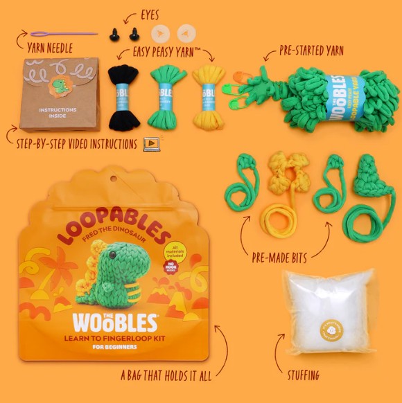 A flat lay of The Woobles Fred the Dino Loopables Kit displays yarn, needle, instructions, plastic eyes, pre-started yarn, pre-made bits, stuffing, and a yellow dinosaur storage bag for fun beginner crafting. Text labels highlight each item.