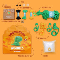 A flat lay of The Woobles Fred the Dino Loopables Kit displays yarn, needle, instructions, plastic eyes, pre-started yarn, pre-made bits, stuffing, and a yellow dinosaur storage bag for fun beginner crafting. Text labels highlight each item.