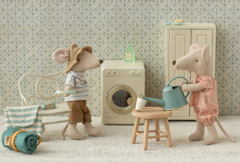 Two Maileg mice enjoy dollhouse play in a cozy laundry room. One, in a hat and striped shirt, sits near a bench; the other wears pink and tends to a plant. In the scene is the Maileg Toothbrush Set In Bag, Mouse - Blue Stripe by Maileg.