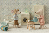 Two Maileg mice enjoy dollhouse play in a cozy laundry room. One, in a hat and striped shirt, sits near a bench; the other wears pink and tends to a plant. In the scene is the Maileg Toothbrush Set In Bag, Mouse - Blue Stripe by Maileg.