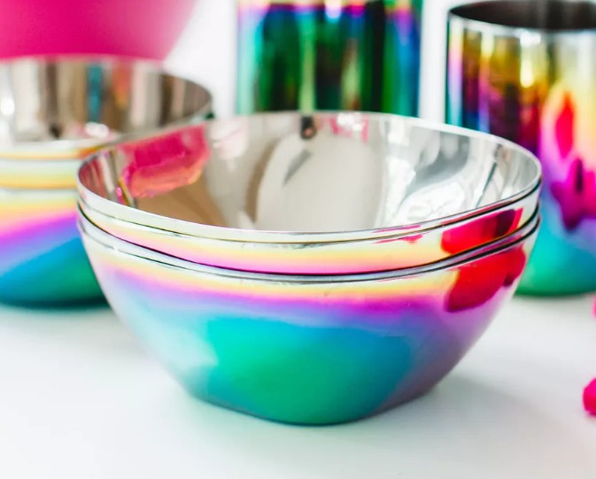 Two shiny ahimsa Rainbow Smart Snacking Bowls (12 oz, pack of 4) with a vibrant, multicolored gradient finish are stacked, while matching colorful kitchenware appears in the background.