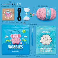 Flat lay of The Woobles Lulu the Elephant Crochet Kit, showing pink yarn, pre-started white yarn, stuffing, crochet hook, eyes, needle, black yarn, embroidered patch, and instructions in a zip bag on a blue background.