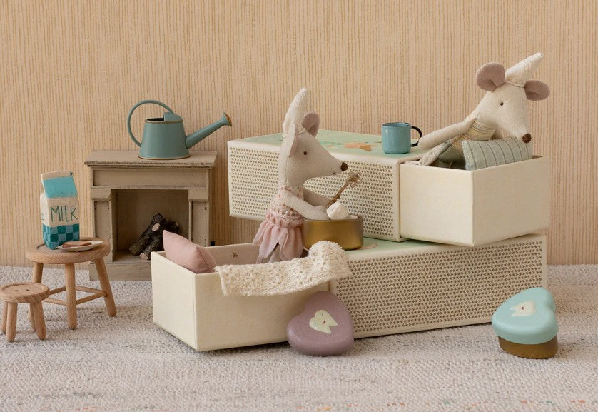 The Maileg Tooth Fairy Mouse, Little Sister in Matchbox by Maileg sits in a cream matchbox bed, surrounded by tiny furniture, a milk carton, watering can, and pastel heart-shaped boxes on a soft rug with a beige background.