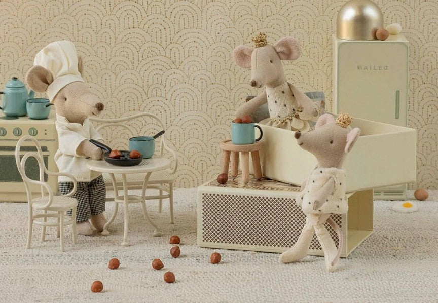 Three Maileg mice gather in a cozy kitchen. One, dressed as a chef, cooks at the table with the Maileg Cafe Set, Mouse, Small - Off White while the others watch. Scattered nuts and tiny appliances complement the soft neutral Maileg decor.