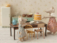 Two Maileg Mouse dolls in dresses stand by a table with a layered cake and plates in a cozy vintage kitchen, complete with the Maileg Fridge, Mouse - Off White, plus a miniature sink and stove for added charm.