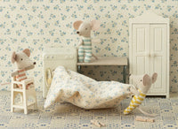 Three Maileg baby mice in striped outfits play in a cozy dollhouse room with a tiled floor and floral wallpaper, interacting with the Maileg High Chair, Mouse - Off White, plus a miniature washing machine, blanket, bench, and wardrobe.