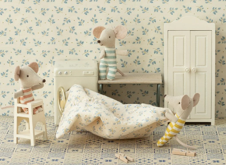 Three Maileg baby mice in striped outfits play in a cozy dollhouse room with a tiled floor and floral wallpaper, interacting with the Maileg High Chair, Mouse - Off White, plus a miniature washing machine, blanket, bench, and wardrobe.