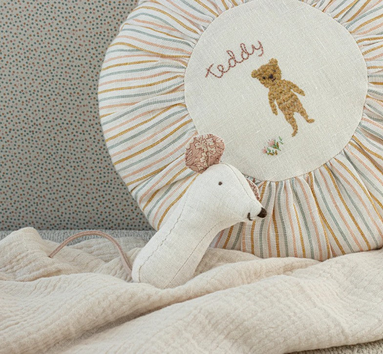 A Maileg Lullaby Friends, Mouse Rattle - Nature by Maileg rests on a cream textured blanket, with a round cushion embroidered with “teddy” and a bear set against a dotted background.