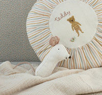 A Maileg Lullaby Friends, Mouse Rattle - Nature by Maileg rests on a cream textured blanket, with a round cushion embroidered with “teddy” and a bear set against a dotted background.