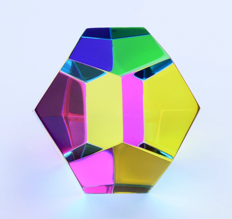 The Mundus by CMY Cubes is a geometric glass sculpture with hexagonal faces that refracts vibrant yellow, pink, green, blue, and purple colors onto a light background.