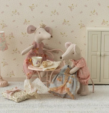 Two Maileg plush mice in dresses enjoy a tea party at a small dollhouse table, perfect for imaginative play. One is the Maileg Big Sister Mouse, elegantly dressed in her Princess dress as they share miniature treats. Tea accessories are set on the table, with a tiny door and lamp adding to the cozy atmosphere.