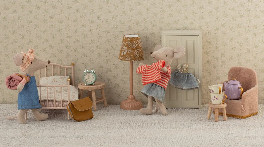 Two Maileg mice relax in a dollhouse room with a bed, lamp, wardrobe, and the Maileg Folding Chair, Mouse - Powder. One mouse carries a rolled blanket and bag; the other holds a red-striped sweater. A table is set for tea—perfect for portable play.