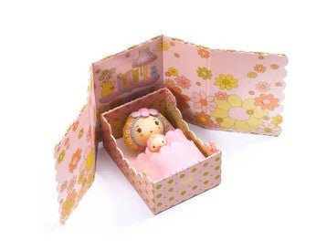 A small DJECO Tinyly Rose doll, wrapped in a pink blanket, lies in a decorated pink and yellow floral box with open flaps featuring charming illustrations and flower patterns—perfect for imaginative play or as an enchanting addition to any DJECO Tinyly Rose Tinyroom miniature playset.