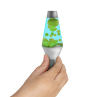 A hand holds the Lava Squish n Flow by Schylling Toys—a small sensory toy featuring a silver base and cap, filled with blue liquid and floating green blobs, designed for stress relief—set against a plain white background.