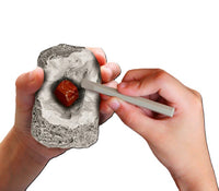 Using a Thames & Kosmos I Dig It! Rocks & Fossils Mini Excavation kit, a person chisels a shiny red gemstone from gray rock, holding the rock in one hand and the chisel in the other, set against a white background.