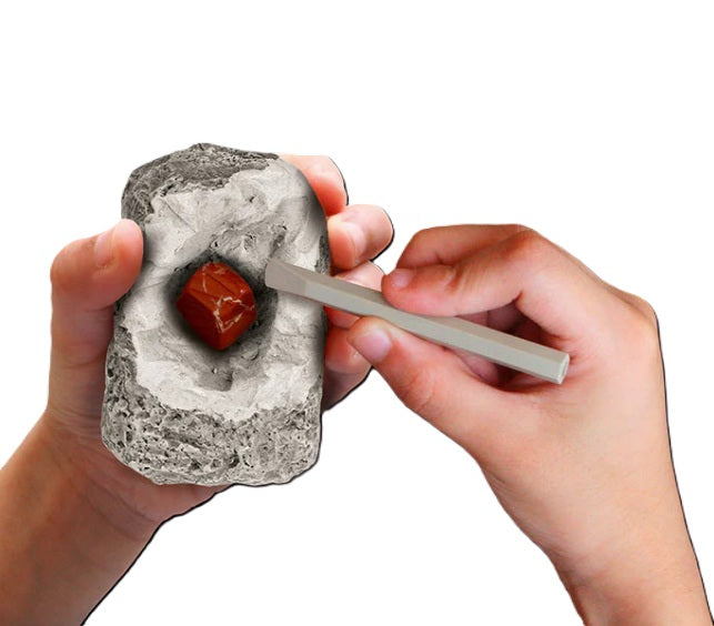 Using a Thames & Kosmos I Dig It! Rocks & Fossils Mini Excavation kit, a person chisels a shiny red gemstone from gray rock, holding the rock in one hand and the chisel in the other, set against a white background.