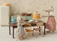 Two Maileg mice in dresses stand by a table with the Maileg Party Cake And Cake Stand, Mouse - Off White, plus plates and teapot, in a miniature kitchen filled with Maileg accessories like a fridge, bowls, and spoons.