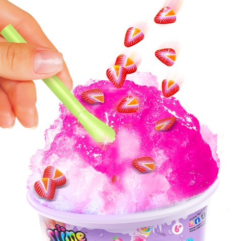 A hand stirs bright pink So Slime Ice Slime Mini Cup by License 2 Play with a green spoon, as heart-shaped strawberry pieces fall onto the slime, creating a moment of sensory fun reminiscent of playing with ice slime or mixing a mini slime cup.