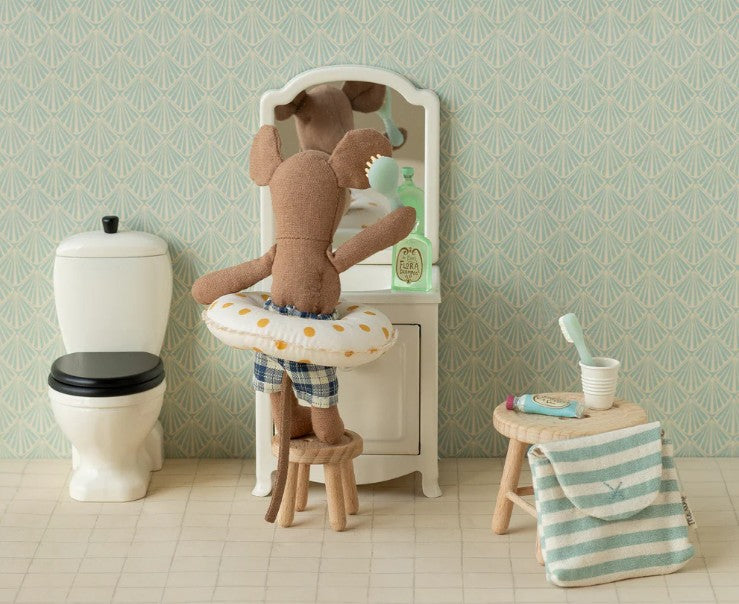 The Maileg Toothbrush Set in Bag, Mouse - Blue Stripe by Maileg features a mouse in plaid shorts with a swim ring, brushing its teeth at a vanity mirror. A toilet, towel, and holder complete this cheerful dollhouse bathroom scene.