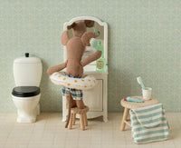 The Maileg Toothbrush Set in Bag, Mouse - Blue Stripe by Maileg features a mouse in plaid shorts with a swim ring, brushing its teeth at a vanity mirror. A toilet, towel, and holder complete this cheerful dollhouse bathroom scene.