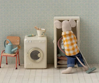 A stuffed mouse in a yellow plaid shirt vacuums beside the Maileg Washing Machine, Mouse – Off white, surrounded by other Maileg furniture and accessories in a charming dollhouse-style room.