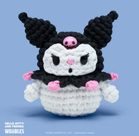 A crocheted Kuromi plush from The Woobles' Kuromi Crochet Kit features a black jester hat with pink skull, black and pink details, and a white body, set against a light blue background—perfect for Kuromi fans.
