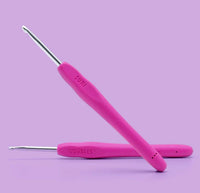 Two pink crochet hooks from The Woobles Suri the Cheetah Crochet Kit, with ergonomic metallic handles for easy grip, are arranged on a purple background—ideal for beginners or making a cute crochet animal.