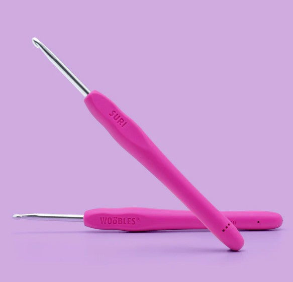Two pink crochet hooks from The Woobles Suri the Cheetah Crochet Kit, with ergonomic metallic handles for easy grip, are arranged on a purple background—ideal for beginners or making a cute crochet animal.