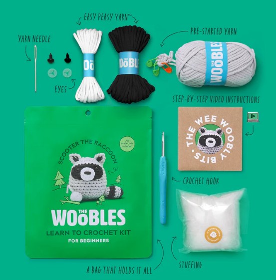 A flat lay of The Woobles Scooter the Raccoon Crochet Kit, with yarn, crochet hook, needle, eyes, stuffing, video instructions, and a green raccoon bag—everything beginners need to make a cute amigurumi raccoon.