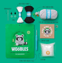 A flat lay of The Woobles Scooter the Raccoon Crochet Kit, with yarn, crochet hook, needle, eyes, stuffing, video instructions, and a green raccoon bag—everything beginners need to make a cute amigurumi raccoon.