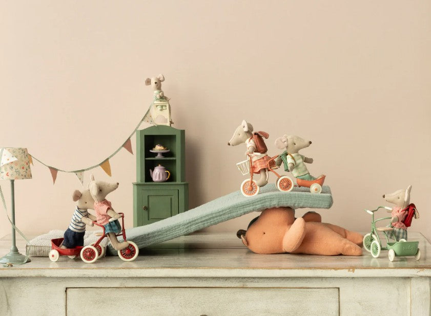 Maileg toy mice pedal on tricycles beside the Maileg Tricycle Garage (Coral), with a small green cabinet, festive lamp, and Mouse tea set adding to the playful Maileg scene.