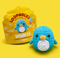 A blue crocheted penguin toy sits before yellow packaging labeled "Pierre the Penguin Loopables Kit" from The Woobles, featuring an image of the same penguin on the box.