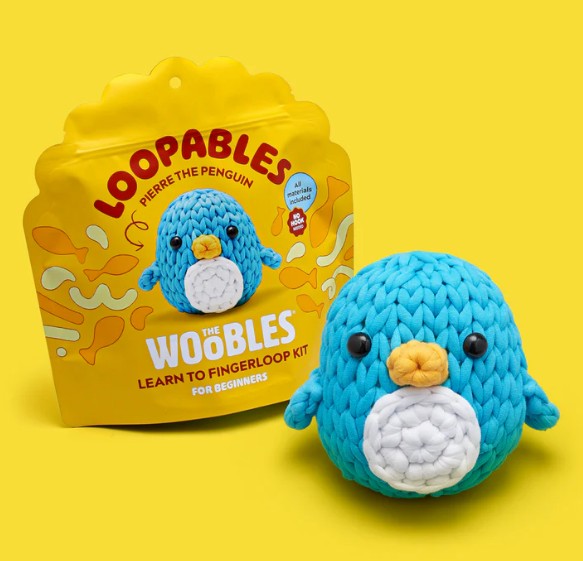A blue crocheted penguin toy sits before yellow packaging labeled "Pierre the Penguin Loopables Kit" from The Woobles, featuring an image of the same penguin on the box.