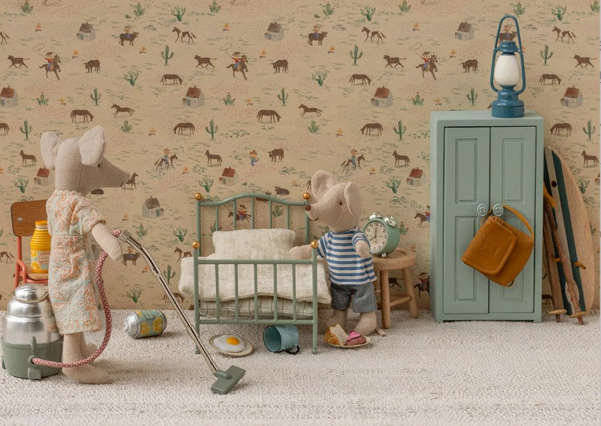 Two Maileg mouse dolls in a tiny room: one vacuums near the Maileg Vintage Single Bed, Mouse - Mint; the other stands by a green wardrobe with a lantern and bag. The room features western-themed wallpaper and mini household items.