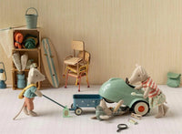 In a playful Maileg scene, three toy mice get a Maileg toy car ready for imaginative adventures in the playroom. The Maileg Driver Mouse pulls a blue wagon, while another inspects under the car and Little Brother Mouse from Maileg checks under the hood. Shelves, chairs, and various toys add to the charming backdrop.