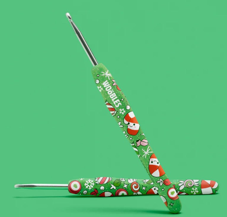 The Woobles Peppi the Candy Cane Crochet Kit includes two green crochet hooks with Santa faces, candy canes, and snowflakes—perfect for beginners or holiday crafting. One hook leans diagonally against the other on a green background.