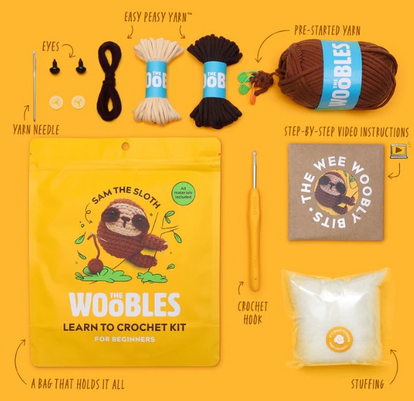 A flat lay of The Woobles Sam the Sloth Crochet Kit for beginners, including yarn, eyes, needle, crochet hook, stuffing, a step-by-step guide, and a yellow bag featuring Sam the cartoon sloth.