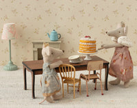 Two Maileg toy mice, featuring Mum Mouse wearing the Maileg Queen dress, stand beside a table arranged with plates and a layered cake; the cozy miniature room scene is decorated with vintage-style furniture, a lamp, and a teapot.