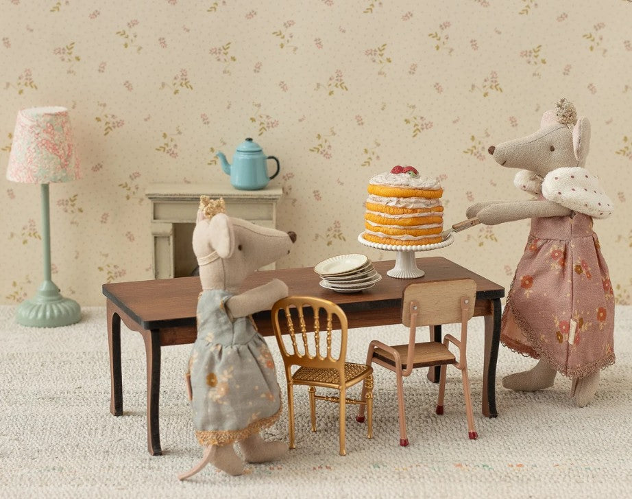 Two Maileg toy mice, featuring Mum Mouse wearing the Maileg Queen dress, stand beside a table arranged with plates and a layered cake; the cozy miniature room scene is decorated with vintage-style furniture, a lamp, and a teapot.