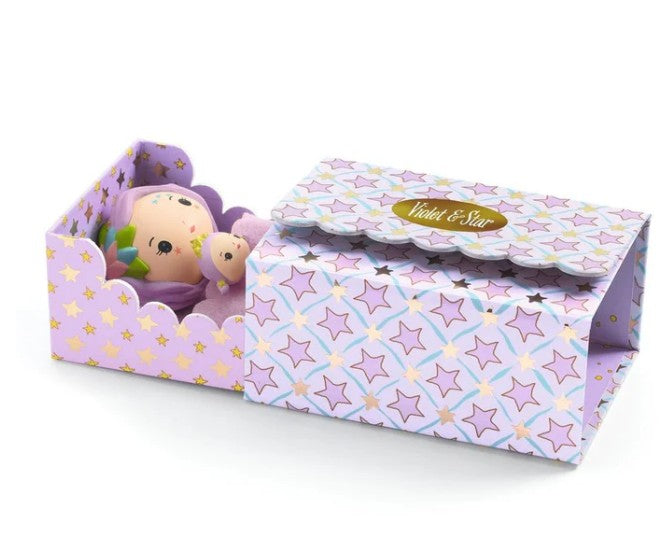 A small DJECO Tinyly Violet Tinyroom doll dressed in a purple outfit rests inside a decorative box with pastel star patterns and a Violet & Star label. The partially open lid reveals the charming doll within—ideal for imaginative play and creating your own miniature room playset from DJECO.