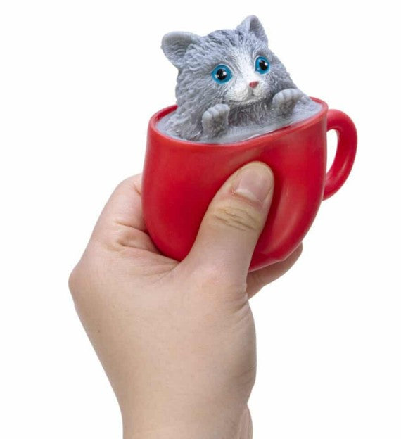 A hand holds a Pop-a-Chino Kittens cup by Schylling Toys, featuring a cute gray kitten figurine playfully popping out of the red cup. This charming cat fidget toy provides soothing stress relief and is delightful to play with. The background is plain white.